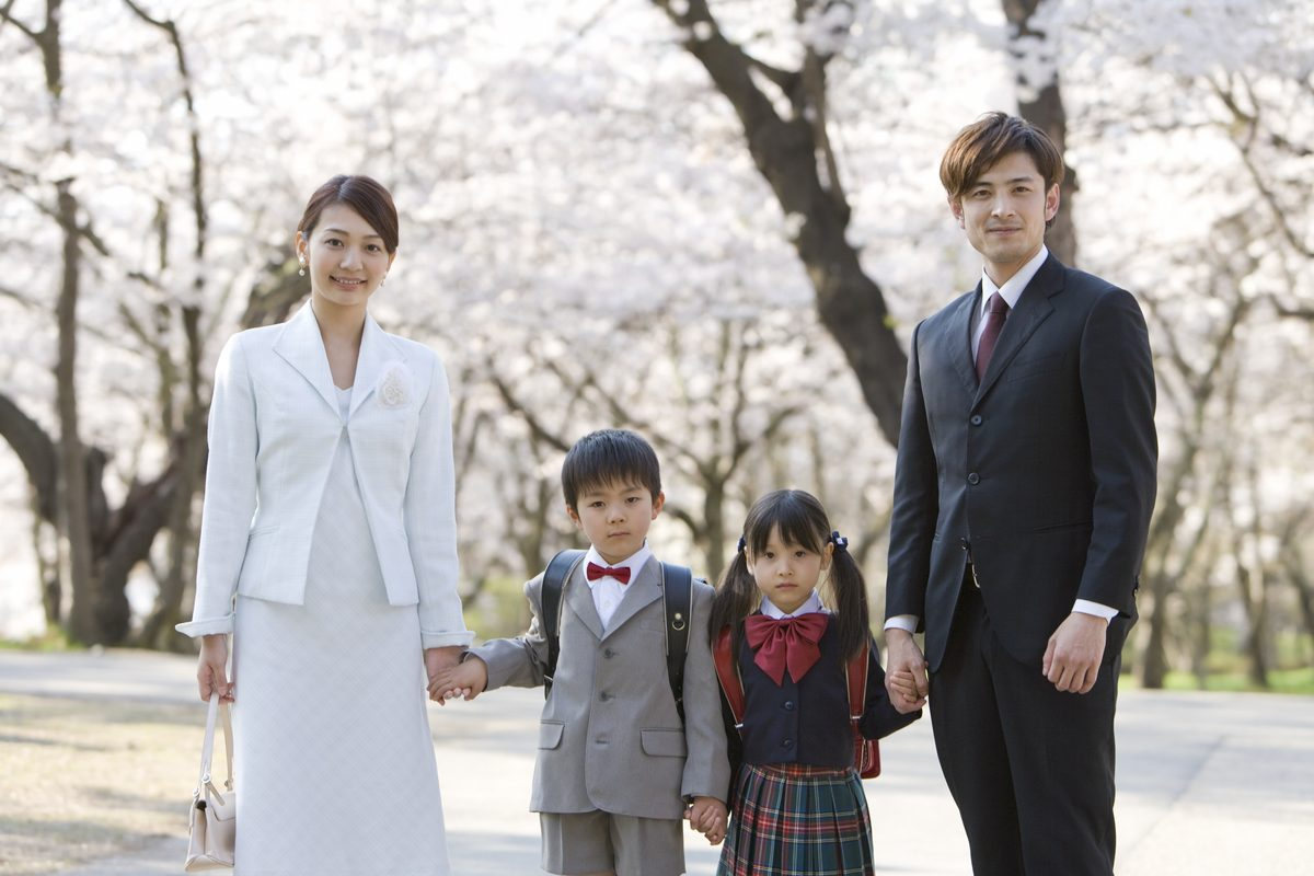 桜並木の下で入学式に向かう家族と、ダークスーツの父親